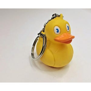 May include: A yellow rubber duck keychain featuring a bright orange beak and white eyes with blue pupils. The duck is attached to a silver-colored metal chain and a circular split ring, providing a functional way to secure keys or other small items.