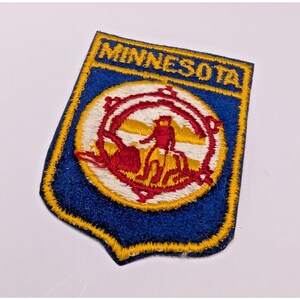 Puede incluir: Un parche bordado en forma de escudo con fondo azul oscuro y borde amarillo. La palabra MINNESOTA está bordada en amarillo en la parte superior. Un diseño circular central muestra una figura roja sobre fondo blanco con detalles amarillos.