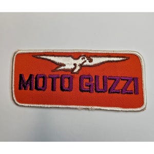 May include: Embroidered Moto Guzzi patch. The patch has an orange background with the words "MOTO GUZZI" in purple. A white bird graphic is above the text. The patch has a white border.