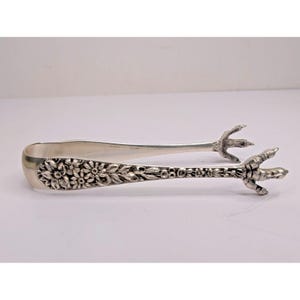 May include: A silver-toned metal serving tong featuring an intricate embossed floral pattern along the handle and arms. The ends of the tongs are shaped like detailed bird talons or claws, designed to grip items securely with a textured finish.