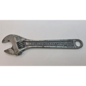 May include: A silver-colored metal adjustable wrench with a knurled adjustment screw. The handle is embossed with the text 4 IN CRESCENT - AT14 - MADE IN U.S.A. The tool features a movable jaw and a circular hole at the end of the handle.