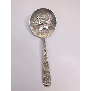 May include: A silver-toned serving spoon featuring an embossed bowl with a detailed cluster of grapes, leaves, and berries. The handle is decorated with an intricate, continuous floral pattern that extends from the bowl to the rounded end.