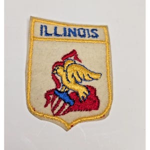 Op de afbeelding: Een schildvormige geborduurde patch met een crèmekleurige vilten achtergrond. Het woord ILLINOIS is bovenaan in blauw geborduurd. Daaronder staat een gouden adelaar afgebeeld op een blauw en rood schild, alles omlijnd met geel borduurgaren.