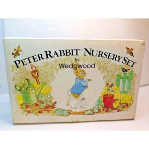 May include: A rectangular white box labeled Peter Rabbit Nursery Set by Wedgwood. The box features a central illustration of Peter Rabbit in a blue jacket, surrounded by garden elements including a green watering can, a shovel, and plants.