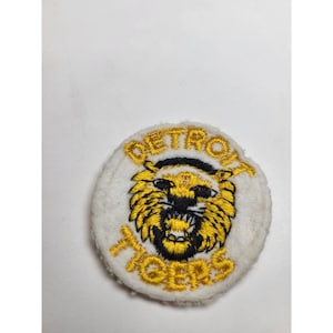 May include: A circular, white patch with gold embroidered text reading "DETROIT TIGERS". The patch features a yellow and black tiger head design, a vintage sports collectible.
