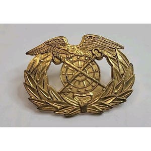 May include: A gold-colored metal emblem featuring an eagle with outstretched wings above a wheel and crossed keys. The wheel is surrounded by a laurel wreath. The emblem has a detailed, vintage appearance.