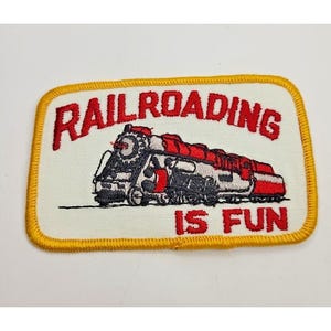 May include: A rectangular embroidered patch with a white background and a yellow border. It features a red and grey steam locomotive illustration in the center. The red embroidered text reads RAILROADING above the train and IS FUN below it.