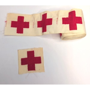 May include: A roll of off-white fabric tape featuring a repeating pattern of red crosses, accompanied by a separate square piece of off-white fabric with a single red cross centered on it. The fabric has a visible woven texture throughout.