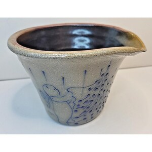 May include: A stoneware pouring bowl with a light gray speckled exterior and a dark brown interior. The exterior features a blue line drawing of a figure holding an umbrella under blue rain dots, with a wide rim and a small integrated pouring spout.