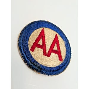 May include: A circular embroidered patch featuring a beige center with the letters AA stitched in red thread. The patch is bordered by a thick, dark blue stitched edge, creating a distinct contrast between the inner circle and the outer rim.