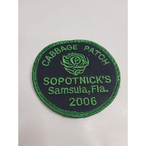 Pode incluir: Um emblema bordado circular com fundo azul-marinho escuro e borda verde brilhante. O emblema apresenta um gráfico de uma couve verde e o texto CABBAGE PATCH, SOPOTNICK'S, Samsula, Fla. e 2006 bordados com fio de cor verde.