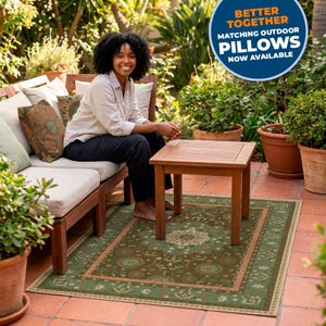 May include: A rectangular outdoor area rug featuring a dark green background with a central floral medallion and a decorative border in shades of tan and cream. The rug is placed on a terracotta tiled patio beneath a wooden side table.