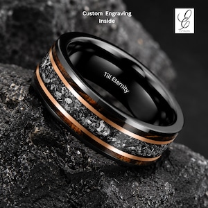 Puede incluir: Un anillo negro con un acabado pulido, con una banda de madera oscura y una incrustación central de material gris moteado. El anillo tiene una fina banda de oro rosa a cada lado de la madera. El texto "Till Eternity" está grabado en el anillo.