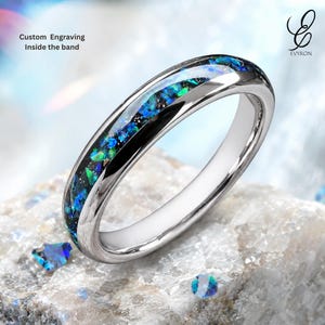 Könnte beinhalten: Ein silberfarbener Ring mit einer zentralen Einlage aus zerkleinertem, blau und grün schillerndem Material. Das Band hat eine polierte metallische Oberfläche. Der Text auf dem Bild lautet: Custom Engraving Inside the band und das EVYRON-Logo.