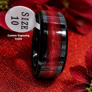 Könnte beinhalten: Ein schwarzer und roter Ring mit einem Größenetikett 10. Der Ring hat ein glänzendes schwarzes Band mit einem roten Mittelteil. Das Etikett lautet "SIZE 10" und "Custom Engraving Inside". Der Ring liegt auf einer roten Oberfläche.