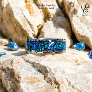 Puede incluir: Un anillo de metal plateado con una incrustación ancha de fragmentos iridiscentes azules y verdes similares al ópalo. La banda tiene un acabado pulido e incluye un pequeño detalle blanco en forma de flecha. El texto dice Custom Engraving Inside the band.