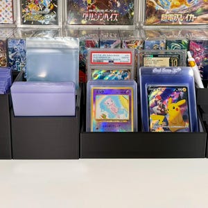 May include: A black modular desk organizer system featuring multiple compartments for trading cards in protective sleeves and hard cases, along with dedicated slots for pens, scissors, and small accessories, arranged in a linear configuration.