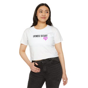 May include: A white short-sleeved crop top featuring the black text "LICENSED TO SLAY" printed on the chest, accompanied by a small, bright pink hand-drawn heart graphic positioned directly below the text on the right side.
