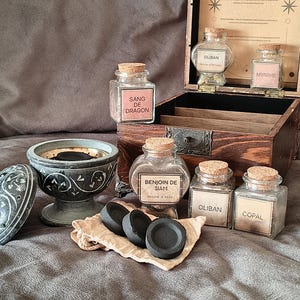 Può includere: Una scatola di legno scuro contenente barattoli di vetro con tappi di sughero etichettati Sang de Dragon, Oliban, Myrrhe, Benjoin de Siam e Copal. Accanto alla scatola c'è un bruciatore di incenso in pietra grigia intagliata con coperchio.