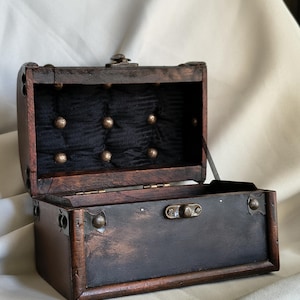 Peut inclure: Un coffre en bois brun foncé avec un couvercle à charnière et un loquet en métal. L'intérieur du couvercle est doublé de tissu noir capitonné avec de petits clous métalliques. Les coins et bords extérieurs sont renforcés par des ferrures.