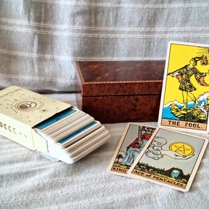 Peut inclure: Un jeu de cartes The Classic Tarot dans une boîte crème avec des illustrations de phases lunaires, une boîte en bois brun foncé et trois cartes de tarot exposées : The Fool, King of Swords et Ace of Pentacles, avec des illustrations colorées.