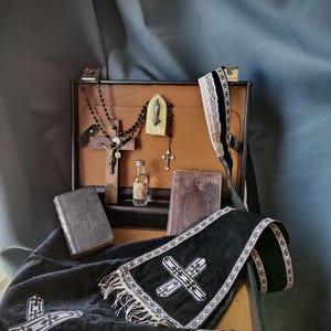 Può includere: Una valigetta foderata in pelle contenente un crocifisso in legno con perline nere, una piccola fiala di vetro, un ciondolo religioso, due libri rilegati in pelle e una stola in velluto nero con croci ricamate bianche e frange.