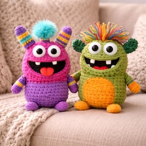 Könnte beinhalten: Zwei handgefertigte Monsterpuppen aus Häkelarbeit. Eine ist rosa und lila mit einem blauen Bommel, die andere ist grün und orange. Beide haben große weiße Augen, offene Münder und kleine Arme und Beine. Sie sitzen auf einer beigen Oberfläche.