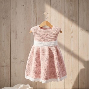 May include: A handmade, light pink crochet dress for a child, hanging on a wooden hanger. The dress features a white waistband and white trim along the hemline. The dress is displayed against a light brown wooden background.