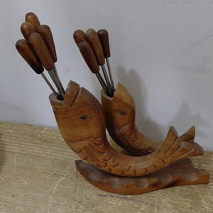 May include: A wooden fish-shaped serving set. Two carved wooden fish hold metal-tipped skewers. The fish are light brown with detailed fins and scales. The skewers have wooden handles. The set is designed for serving.