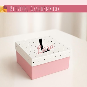Könnte beinhalten: Eine quadratische Geschenkbox mit einem einfarbig rosa Unterteil und einem weißen Deckel mit schwarzem Punktmuster. Der Deckel ist mit einem großen schwarzen Buchstaben L und dem Namen Lara in rosa Schreibschrift personalisiert.