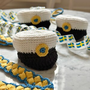 May include: Handmade crocheted graduation caps in white, black, and yellow, with a blue and yellow ribbon. The caps have a circular yellow and blue emblem. The ribbon features the Swedish flag. A charming, handcrafted design.