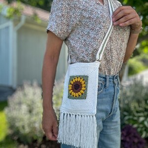 May include: White crochet crossbody bag with a sunflower design and fringe detail. The sunflower is made with brown, yellow, and green yarn. The strap is white with a green stripe. The bag is worn over the shoulder.
