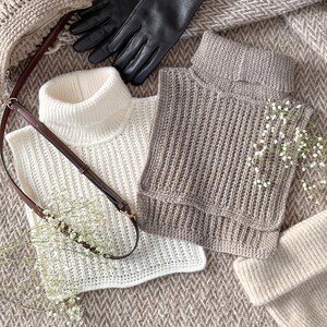 May include: An assortment of winter accessories, including a cream-colored knitted turtleneck vest, a gray ribbed knit vest, black leather gloves, a beige handbag, and a matching beanie. The items are arranged on a textured, neutral-toned fabric.