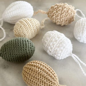 May include: A collection of handmade crochet Easter egg ornaments in various colors, including white, beige, and olive green. Each egg has a textured surface and a small loop for hanging. The eggs are arranged on a light-colored surface.