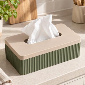 May include: A rectangular tissue box holder featuring a matte beige lid and a dark olive green base with a vertical ribbed texture. A single white tissue is pulled through the central oval opening on the top of the container.