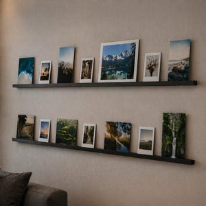 Op de afbeelding: Twee zwarte zwevende planken aan een beige muur tonen ingelijste landschapsfoto's. De foto's variëren in grootte en tonen natuursferen, waaronder bergen, water en bomen. De planken zijn boven een grijze bank gemonteerd.