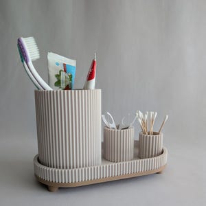 Puede incluir: Un conjunto organizador de baño de color beige con una textura acanalada en el exterior. El set incluye un soporte cilíndrico grande, dos soportes cilíndricos más pequeños y una base de bandeja ovalada con patas pequeñas.