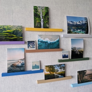 May include: A collection of ten rectangular wall-mounted floating shelves in various colors including yellow, purple, orange, blue, beige, white, green, and light blue, each displaying a different landscape or portrait photograph on a textured wall.