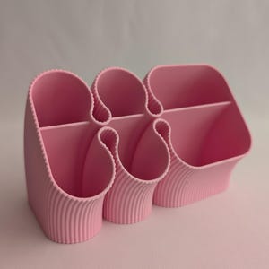 May include: A light pink desk organiser featuring a series of interconnected, vertical compartments with a ribbed, textured exterior. The structure includes multiple open-top sections of varying heights and shapes designed for holding stationery items.