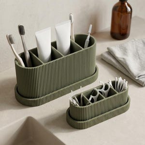 Puede incluir: Dos organizadores de baño de color verde oliva con textura acanalada vertical y bandejas a juego. El organizador más grande contiene cepillos de dientes y tubos de pasta dental, mientras que el pequeño tiene hilo dental y bastoncillos.