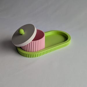 Pode incluir: Uma bandeja oval verde-limão com borda canelada e um recipiente cilíndrico rosa correspondente com exterior canelado. O recipiente tem uma tampa circular cinza-claro com um pequeno botão esférico verde-limão, pousada entreaberta.