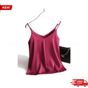 May include: A burgundy camisole with a V-neck and thin straps hangs on a white hanger. A black quilted handbag with a chain strap is to the right. The word "NEW" is in a red box in the upper left corner.