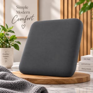 May include: A square, dark gray, smooth-textured cushion with rounded corners, positioned upright on a circular wooden base. The cushion features a solid, matte finish and a firm, uniform appearance, providing a neutral and clean aesthetic element.