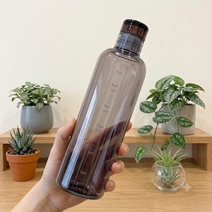 May include: A translucent dark gray plastic water bottle with a black screw-top lid. The bottle features white printed time markers ranging from 8am to 5pm, designed to track hydration throughout the day, with a clear cylindrical body shape.