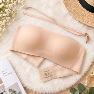 May include: A nude-colored strapless bandeau bra with a smooth, seamless finish. The garment features a back closure with hook-and-eye fasteners and includes a detachable, adjustable thin strap. The interior label indicates size M, nylon/spandex blend.
