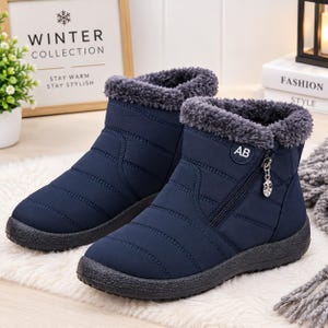 May include: A pair of navy blue ankle boots featuring a quilted exterior, a dark gray faux fur lining at the collar, and a side zipper closure with a silver-toned leaf-shaped charm. The boots have a black textured rubber sole and the letters AB.