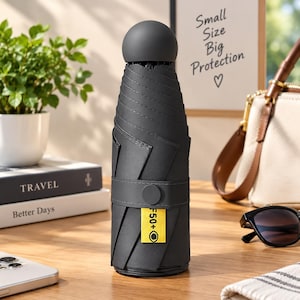 Puede incluir: Un paraguas plegable de color negro mate compacto con un mango redondeado. Una correa horizontal asegura la cubierta plegada, con una etiqueta rectangular de color amarillo brillante impresa con el texto UPF50+ y un icono circular.