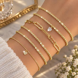 May include: A set of six gold-toned bracelets featuring various designs, including thin textured chains, a link chain, a single round clear stone charm, and a small heart-shaped charm encrusted with tiny clear stones, all worn together.