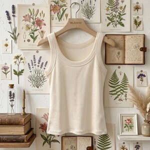 May include: A cream-colored ribbed tank top with a wide scoop neckline and thick shoulder straps, hanging on a wooden hanger labeled BLSSNZ. The garment features a simple, sleeveless design with a clean hemline and a soft, textured fabric finish.
