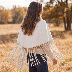 May include: A cream-colored poncho featuring a V-shaped yoke with decorative whipstitching along the seam. The garment has long, thin fringe detailing hanging from the bottom edge, creating a layered appearance over a beige sweater and dark jeans.